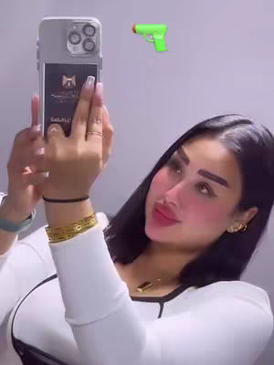 A woman is holding a smartphone, taking a selfie. A cartoonish green gun is floating above her head. She has long black hair, and is wearing heavy makeup. She's wearing a white shirt and gold jewelry.