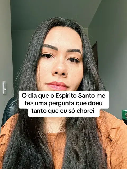A woman with long dark hair is centered in the frame, looking directly at the camera with a serious expression. A white rectangular text box overlay covers part of her face, containing text in Portuguese. The background is a muted green wall and a hint of a dark chair.