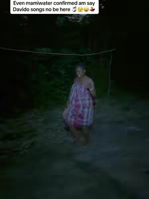 A person, wrapped in a patterned cloth, stands in a dark environment. The background appears to be a field or outdoor space, barely visible in the dim lighting.