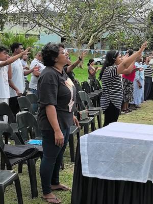 A group of people standing outdoors with their hands raised towards the sky, suggesting prayer or worship. The scene takes place on a grassy area with trees and some buildings in the background. A table covered in white cloth in the foreground.