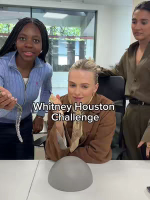 Three women are in an office setting. The central figure sits at a white table in the foreground, with a gray rounded object in front of her. She's wearing a blazer and a patterned scarf. The woman on the left has her hair braided and wears a blue zip-up. The woman on the right is standing wearing a collared shirt. The background includes windows and cabinet.