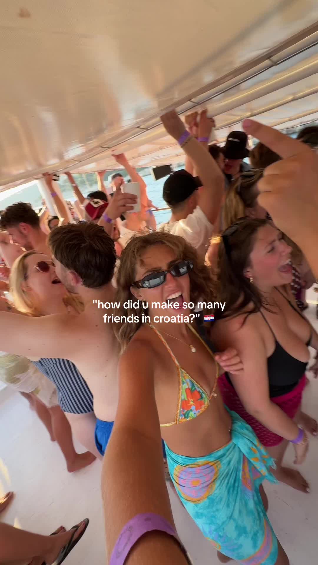 A selfie-style video with a group of people on a boat, celebrating and dancing. Several individuals are in the frame, with one woman in focus, wearing sunglasses and a bikini top. The background shows more people partying and the water. The lighting suggests a sunny day and a vibrant party atmosphere.