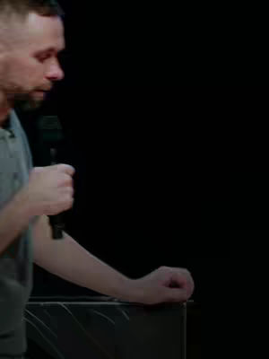 A man standing at a podium or similar structure, seen from a side profile. He holds a microphone in his right hand, his left arm resting on the podium. The background is solid black.