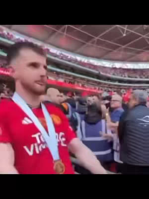 A man in a red sports jersey with a medal around his neck, standing in a stadium filled with people. The background shows a stadium with red seats and a white roof.