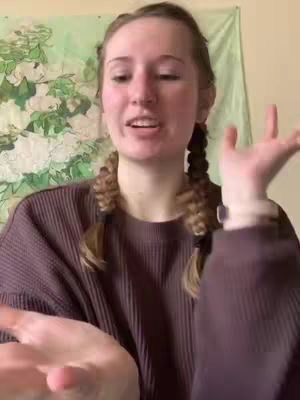 A young woman with long, light-colored hair styled in two side braids is centered in a medium shot. She is looking at the camera and talking, with her right hand raised and open, as if gesturing. She is wearing a brown long-sleeved shirt. Behind her is a light green tapestry with a floral design. The background is otherwise plain, and the lighting appears natural.