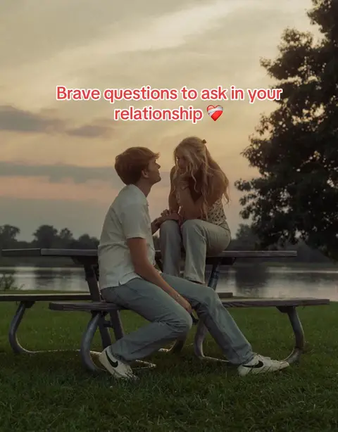 A couple sits on a park bench facing each other, the man looking up at the woman. Sky has sunset colors, with green grass and water in the background.