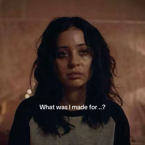 A close-up of a young woman with dark hair and a somber expression. She is looking directly at the camera, with visible tears or moisture around her eyes. The background is a soft, blurred mix of warm colors. Text overlayed at the bottom says, "What was I made for..?"
