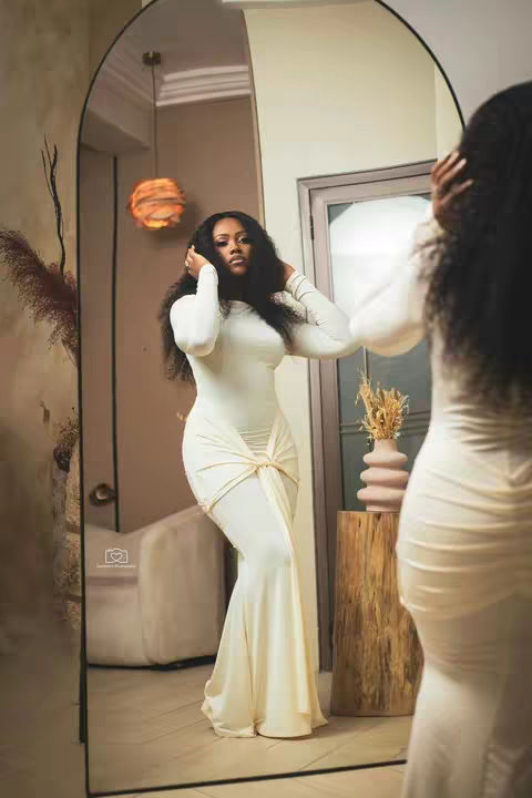 A woman in a white dress poses in front of a large mirror. Her reflection is visible. She's touching her hair and the background appears to be a well-lit room with some decorations.