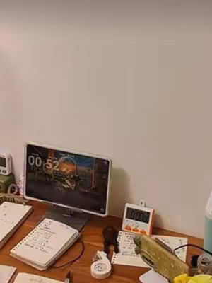A desk setup with a computer screen displaying what appears to be a timer. Several notebooks, a timer, a desk lamp, and other office supplies are scattered across the surface, suggesting a study environment.