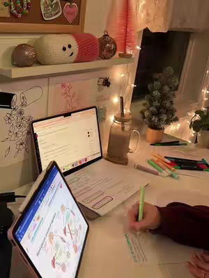 A well-lit desk setup for studying, featuring a laptop and tablet displaying educational content, with a hand taking notes on paper. The setting has warm lighting, a coffee cup, and decorative touches.