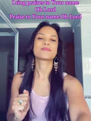 A woman with long dark hair and dangling earrings is looking upward. Text overlays in the image read, "I sing praises to Your name Oh Lord, Praise to Your name Oh Lord". The woman wears a pink top, and has on a ring on her left hand. The setting appears to be indoors, with a glimpse of a window/door in the background.