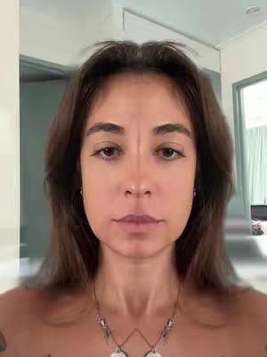 The image is a symmetrical portrait of a person with brown hair and light skin. The person is looking directly at the camera with a neutral expression. The background is out of focus but appears to be an interior space with neutral-colored walls and some visible architectural elements. The color palette is muted, giving the image a natural, realistic feel.