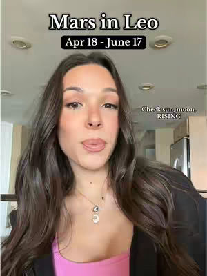 A woman with long dark hair is centered in the frame, looking at the camera with a slight smile and her tongue out. Above her head are the words "Mars in Leo" and "Apr 18 - June 17" in black and white text. The background has a warm-toned interior with visible ceiling lights.