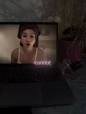 A laptop is open, displaying a video of a woman with a towel wrapped around her head. The screen shows her talking with an open mouth in what seems to be a bathroom setting. Text overlayed on the laptop screen reads 'cannot.' The laptop is on a dark surface, with a perfume bottle and a plant in the background.