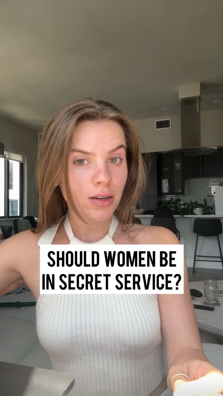 A woman with light skin and long, light brown hair is centered in a well-lit interior. She is wearing a white halter top. A white rectangular text box is positioned on the lower part of her face, overlapping her mouth. Behind her, a modern kitchen is somewhat visible, with a stainless steel range hood and what appears to be a bar area.