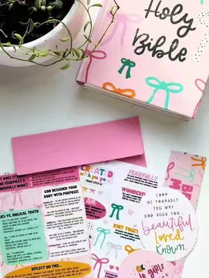 A top-down, neatly arranged composition of a pink covered Holy Bible, surrounded by study papers and notes with colorful illustrations and text. A small potted plant with green foliage rests in the upper left corner, and the items are set against a white surface, conveying a message of faith and study.