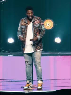 The image shows a man on a stage in front of a purple wall with stage lighting. He is dressed in a casual, yet stylish outfit consisting of a white t-shirt, a plaid jacket, and ripped jeans. He's wearing orange and black sneakers and has dark hair and a beard. The man is gesturing with his hands, likely speaking or presenting. The stage has a reflective surface and a circular light fixture.