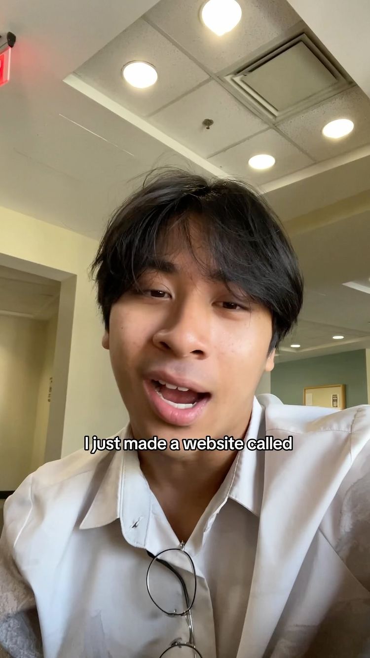 A person with short black hair and glasses is in front of the camera. In the background, there is a white ceiling with lights, and it appears to be a public building setting.