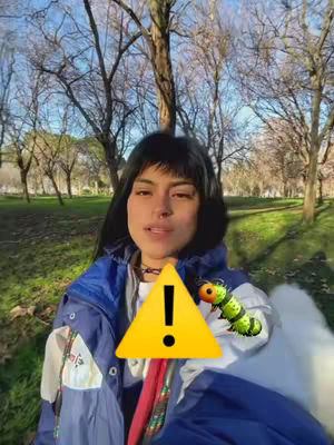A woman is in the foreground of a park-like setting. She is wearing a jacket with a blue and white color scheme. The background features trees without leaves and a blue sky. An alert symbol and a cartoon caterpillar are overlaid on the image.