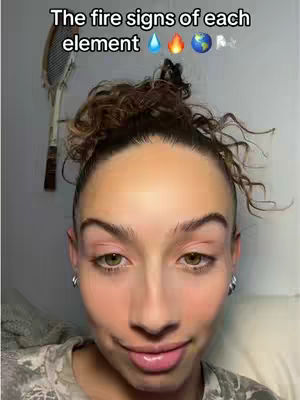 A close-up selfie of a woman with brown curly hair pulled back in a bun, pink-toned makeup, and a slightly smiling expression. Text is overlaid at the top with the words 'The fire signs of each element' followed by emojis of a water droplet, a flame, a globe, and a few indistinct symbols.