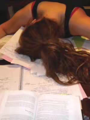 A woman lies face down on a surface covered with papers and notebooks, with long, light brown hair partially obscuring her face. She is wearing black underwear with a red trim. An open book is visible in the foreground.