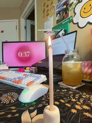 A well-lit desk with a computer monitor displaying a pink screen with the time 0:37. A lit candle stands in the foreground, with a keyboard, mouse, and a glass of iced tea on the desk, all set against a backdrop of various desk items.