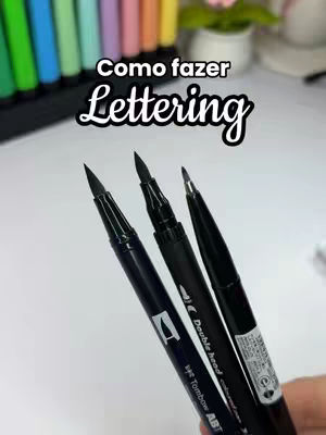 The image shows three black pens angled towards the viewer. The pens have various markings and brand names such as "Tombow ABT". Above the pens, white text on a black background says "Como fazer Lettering". In the background, there is a blurred array of colored markers and a touch of a floral arrangement.