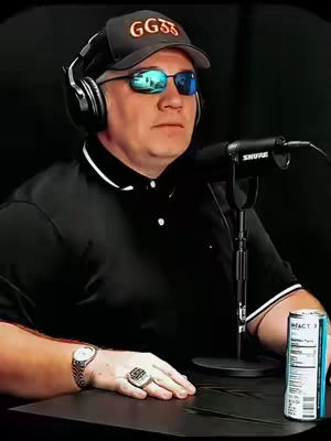 A man wearing sunglasses, a hat, and headphones sits in front of a microphone with a drink in front of him.