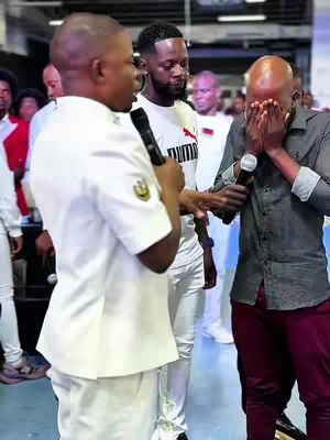 A group of men are in a large indoor space, possibly an event. One man in a white uniform with a microphone is speaking. Another man covers his face with both hands, appearing distressed. Other people are in the background.