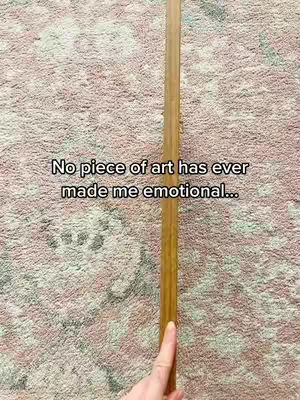 The image shows a close-up of a rug with a wooden rod in the center. The rod is held vertically, with a finger touching it near the bottom. Text overlay reads 'No piece of art has ever made me emotional...'