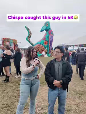 A woman with long brown hair, wearing a white top and light blue jeans, is holding a microphone and interviewing a man wearing glasses, a black jacket and jeans. They are standing in an outdoor setting with a colorful sculpture in the background and other people.