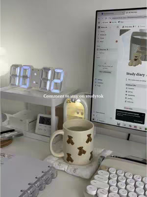 An interior shot of a desk setup. A digital clock displaying the time, a computer monitor displaying a webpage, a mug with bear designs, a keyboard, and a notepad. The overall aesthetic is minimalistic and cozy.