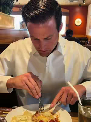 A man is seated at a restaurant table, looking down at a plate of food he is cutting. He is wearing a white dress shirt, and the setting appears to be a casual dining establishment. Soft lighting and a warm color palette dominate the scene.