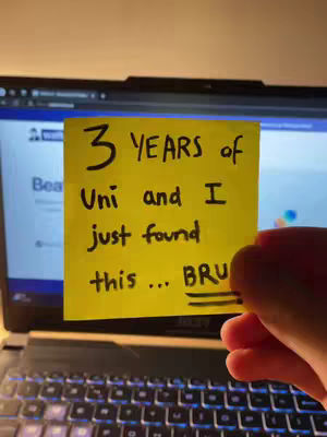 A laptop with an open screen, a yellow sticky note with handwritten text in black marker is held in front of the screen. The hand holding the note is visible. The background suggests a dimly lit environment with warm tones.