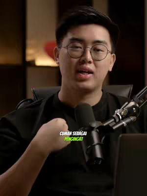A man in glasses speaks into a microphone. Text overlay reads "CUMAN SEBAGAI PENGINGAT" in a yellow-green box. The background is dark, with hints of a blurred interior. The man has short, dark hair and is wearing a black shirt.