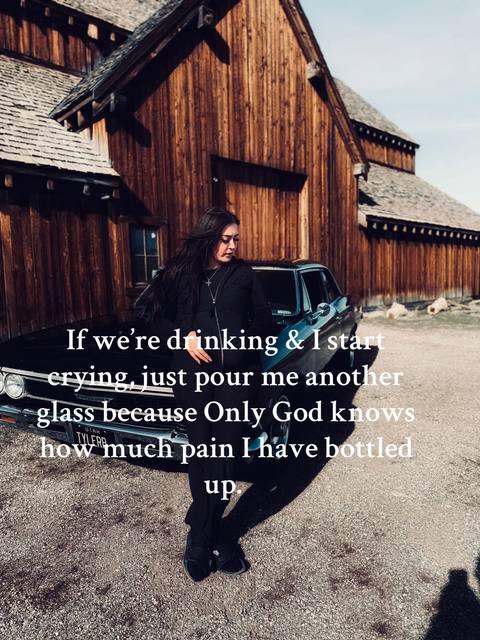 The image shows a woman standing next to a black car, set against a backdrop of a rustic wooden building during a sunny day. Text is overlaid on the image, expressing a feeling of suppressed pain. The woman is wearing a black outfit, with her hair flowing, and with a cross-neckless, completing the composition.
