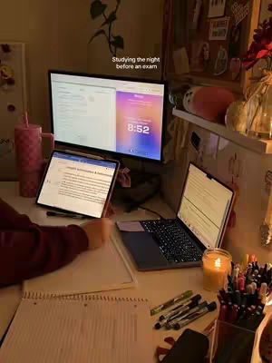 A dimly lit desk with study materials. A laptop, tablet, and computer monitor display study notes. There is a candle lit for ambiance, pens, and a tumbler of pink liquid, implying a study session.