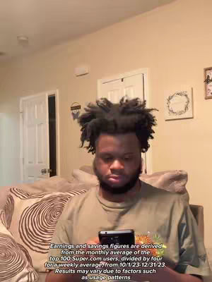 A man with dark, curly hair and a beard is seated, holding a smartphone. He wears a casual olive-green shirt. The backdrop includes a neutral-toned wall with a white door and wall decor, suggesting an interior setting. A section of a sofa with patterned pillows is partially visible.