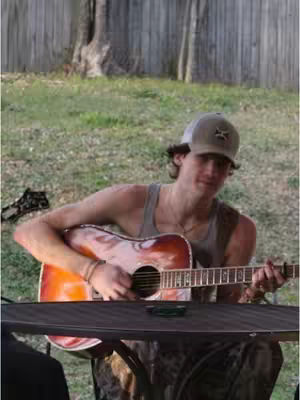 A young man with a guitar sits at a table in an outdoor setting. He wears a baseball cap, tank top, and has a guitar in his hands. The image has a warm tone with natural lighting.