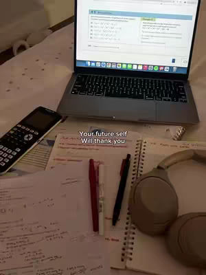 The image is a top-down view of a study setup on a bed. A laptop displaying a screen with what appears to be math problems is at the top, with a calculator and notes below. Pens, a notebook, and headphones are visible, suggesting a study session. The overall composition creates a workspace environment, and the lighting is natural.