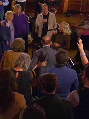 The image captures a stage setting with a diverse group of people, likely during a musical performance or church service. Several individuals are the focus: a blonde woman with a microphone, a man with a microphone singing in front of the crowd. The overall lighting appears warm, and the composition focuses on human connection and expressive gestures like hands raised in worship.