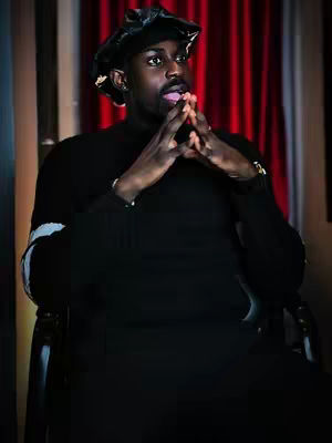 A dark-skinned man sits in a chair, wearing a black turtleneck and hat. His hands are clasped near his face, and he is gazing thoughtfully to the side with his tongue slightly out. The background includes red curtains and a window, suggesting an interior setting.