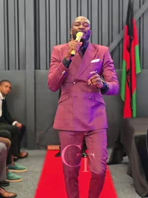 A man in a maroon suit stands on a red carpet, holding a microphone. The background has a dark wall and a flag on a pole. Other people are seated.