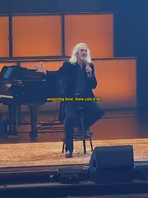 The image shows a man in a concert setting, holding a microphone and singing. There is a piano to the left and a speaker in the foreground.