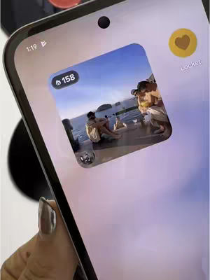 A close-up shot of a smartphone displaying the Locket application. The phone is held in a person's hand, with a silver nail visible, against a blurred background. A square widget from the app shows a picture with people on a beach. The interface elements are displayed, like battery and time on the top left and application features.