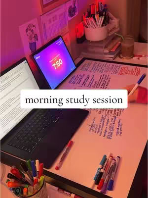 A top-down shot of a study setup featuring two laptops, a tablet with a timer, and papers with handwritten notes. Colored markers and a drink are also visible. The background is lit with pink and purple light.