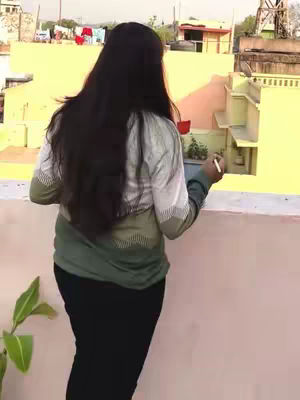 A woman with long dark hair is standing on what appears to be a rooftop or balcony, looking away from the camera towards a yellow building. She is holding a white object and writing with a pen, and the background is filled with buildings.