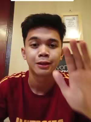 The image shows a young man looking directly at the camera with a slightly open mouth, as if he's about to speak. He's wearing a dark red shirt with yellow stripes on the shoulders and a logo on the chest. His right hand is raised, appearing to be waving. In the background, there's a framed picture on the wall and a partially visible dark brown door or panel to the left. The lighting suggests a well-lit indoor environment.
