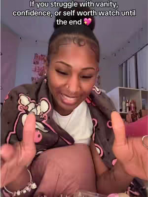 A woman with dark skin and curly black hair pulled into a bun is centered in a selfie-style video. The text overlayed on the image asks viewers to watch the video if they struggle with vanity, confidence, or self-worth. She is wearing a white top and a patterned jacket, and her hands are up, gesturing towards the camera. Behind her, soft lighting and a neutral background hint at an indoor setting.