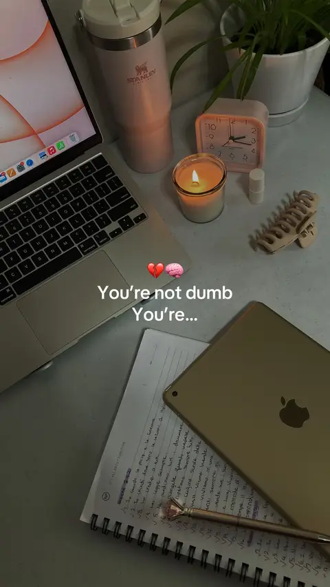 A well-lit, aesthetically arranged desk scene. A laptop is open, next to a pink Stanley tumbler and a lit candle in a clear glass jar. To the right, there is a small pink alarm clock and an open notebook with handwritten notes and a pen, as well as a golden iPad and a hair clip. The overall atmosphere is cozy and focused, with a soft, warm color scheme.
